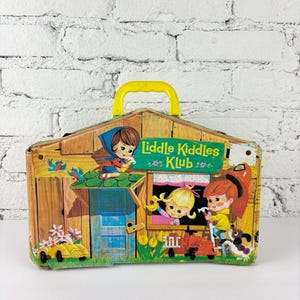 May include: A vintage Liddle Kiddles Klub metal lunchbox featuring a yellow plastic handle and colorful illustrations of cartoon characters, a small house, a yellow car, and a dog on a green lawn against a wooden structure background.