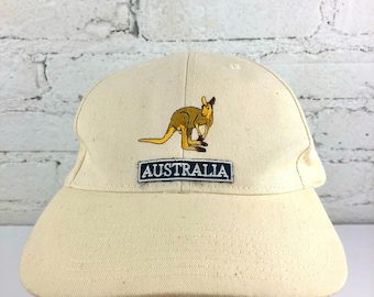 May include: A cream-colored baseball cap featuring an embroidered kangaroo and the word "AUSTRALIA" on a rectangular patch. The cap has a curved brim and a classic six-panel design, suitable for casual wear.