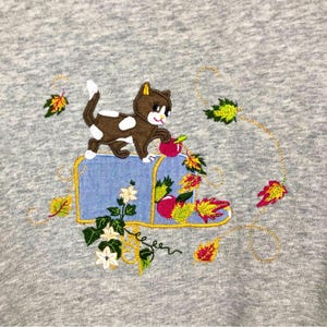 May include: Embroidered grey sweatshirt featuring a brown and white cat on a blue mailbox. The mailbox is decorated with red apples, autumn leaves, and white flowers. The design is accented with gold thread.