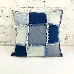 Op de afbeelding: Vierkant denim patchwork kussen in blauwe en lichtgrijze tinten. Het kussen heeft een rasterpatroon met gerafelde randen, wat een textuur en handgemaakte uitstraling creëert. Het kussen is een decoratief woonaccessoire.