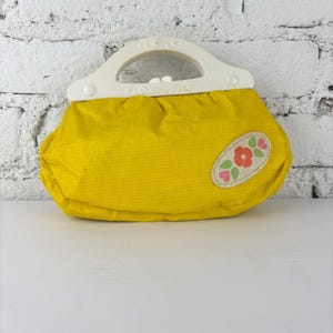 Op de afbeelding: Een vintage gele Fisher-Price handtas met een witte handgreep. De tas heeft een bloemenapplicatie met rode en roze bloemen en groene bladeren. De handgreep is wit met het Fisher-Price logo.
