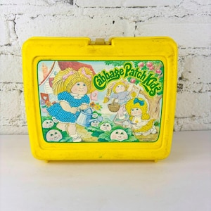 Op de afbeelding: Een gele plastic Cabbage Patch Kids lunchbox. De voorkant heeft een kleurrijke illustratie van Cabbage Patch Kids-personages in een tuin, met de woorden "Cabbage Patch Kids" in groene letters.