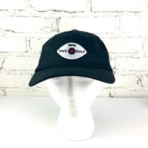 May include: Black baseball cap with a curved brim. The cap features an embroidered white oval patch with the words "MFM FAN CULT" and a red bullseye graphic. The cap is displayed on a white mannequin head.