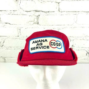 Puede incluir: Un gorro de invierno rojo con orejeras. El gorro presenta un parche rectangular con las palabras "AMANA AG SERVICE COOP" en blanco y azul. El gorro se exhibe sobre una cabeza de maniquí blanca contra una pared de ladrillos blancos.