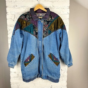 Puede incluir: Chaqueta vaquera vintage con diseño patchwork. La chaqueta presenta un cuerpo de mezclilla azul claro con cuello y canesú estampados en tonos morado, verde azulado y dorado. Dos bolsillos delanteros y cierre de botones completan el diseño.