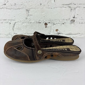 May include: A pair of brown leather clogs with a strap and cut-out design. The clogs have a plaid insole and a rubber sole. The brand name "Mudd" is visible on the insole. The shoes are in good condition.