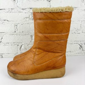 Puede incluir: Un par de botas de invierno marrones vintage. Las botas tienen un diseño acolchado con un ribete de piel sintética en la parte superior. Las suelas son de color marrón claro. Las botas miden aproximadamente 25 cm de alto.