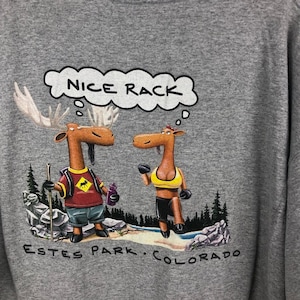 Puede incluir: Una camiseta gris con una imagen de dibujos animados de dos alces. El alce macho lleva una camisa roja y lleva un bastón de senderismo. La alce hembra lleva un sujetador deportivo amarillo y pantalones cortos negros. El texto "NICE RACK" está en una burbuja de pensamiento sobre el alce macho. El texto "ESTES PARK. COLORADO" está en la parte inferior de la camiseta.