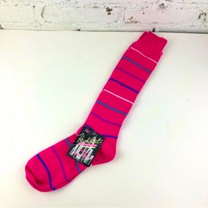 Könnte beinhalten: Ein einzelner, leuchtend pinkfarbener kniehoher Strumpf mit horizontalen Streifen in Blau, Weiß und Türkis. Die Socke hat ein Etikett mit der Aufschrift "High Art" in einer stilisierten Schriftart. Die Socke liegt auf einer weißen Oberfläche.