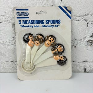 May include: A set of five measuring spoons in a package. The spoons are white with monkey-shaped handles. The packaging reads "5 Measuring Spoons" and "Monkey see...Monkey do."