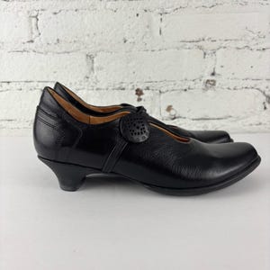 May include: A pair of black leather Mary Jane shoes. The shoes feature a low heel, a rounded toe, and a strap with a decorative circular button. The shoes are set against a white brick wall.