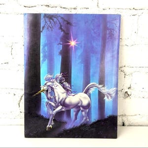 May include: A fantasy art print featuring a white unicorn with a golden horn, set in a mystical forest with tall, dark trees and a glowing star. The unicorn has a flowing mane and tail, with purple accents. The background is a gradient of blues and purples.
