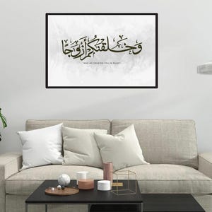 May include: A framed artwork with Arabic calligraphy in black, set against a light gray background. The text reads "AND WE CREATED YOU IN PAIRS." The artwork is displayed above a beige sofa with white pillows, a black coffee table, and a patterned rug.
