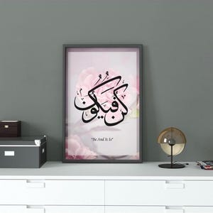 May include: Framed art print with Arabic calligraphy in black on a pink floral background. The print is displayed on a white cabinet with a small wooden box, a gray storage box, and a decorative lamp.