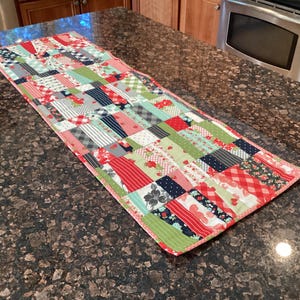 May include: A rectangular patchwork table runner with a variety of fabric patterns in red, green, gray, and white. The runner features floral, striped, and checkered designs, and is edged with a red border. It is placed on a dark brown countertop.