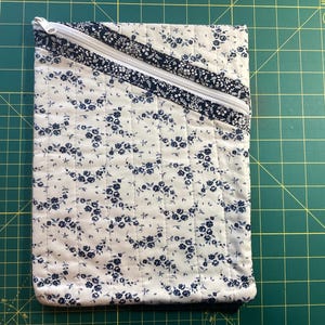 May include: Quilted fabric case with a white background and a pattern of small blue flowers. The case has a diagonal zipper and is trimmed with a dark blue floral fabric. The stitching is visible, creating a textured effect.