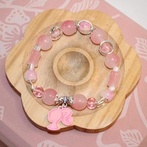 Op de afbeelding: Een roze kralenarmband met een Barbie-silhouetbedel. De armband heeft verschillende roze kralen, waaronder ronde en cilindrische vormen, en zilverkleurige accenten. De armband wordt gepresenteerd op een houten, bloemvormig dienblad.