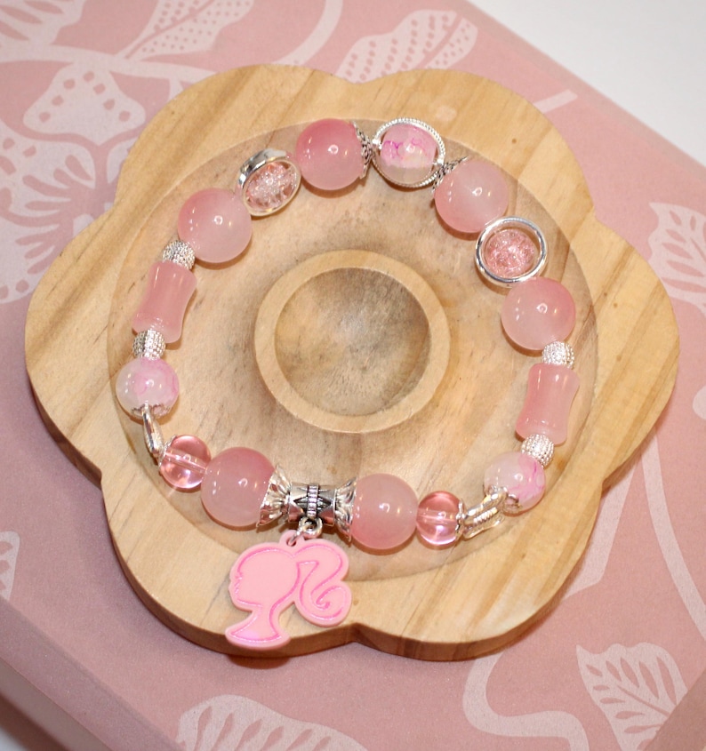 Op de afbeelding: Een roze kralenarmband met een Barbie-silhouetbedel. De armband heeft verschillende roze kralen, waaronder ronde en cilindrische vormen, en zilverkleurige accenten. De armband wordt gepresenteerd op een houten, bloemvormig dienblad.