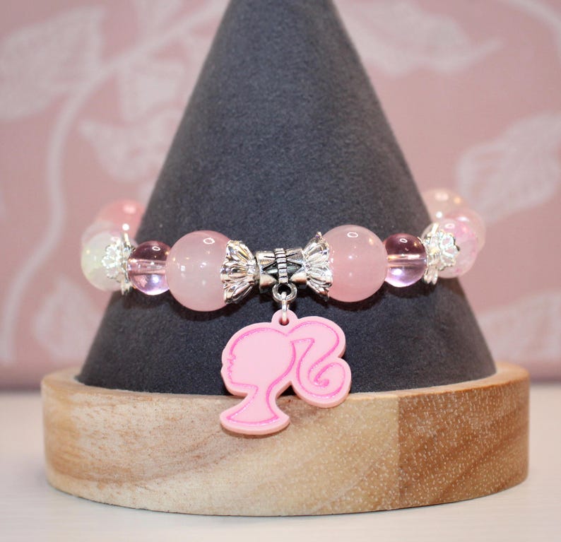 Op de afbeelding: Een roze kralenarmband met een bedel van een vrouwensilhouet. De armband heeft roze en heldere kralen, zilverkleurige afstandhouders en een roze bedel. De armband wordt getoond op een grijze kegelvormige standaard met een houten basis.