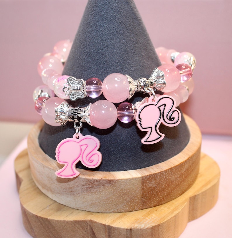 Op de afbeelding: Twee roze kralenarmbanden met zilveren accenten en roze bedels. De armbanden worden gepresenteerd op een grijze, kegelvormige standaard. De bedels hebben de vorm van een vrouwensilhouet.