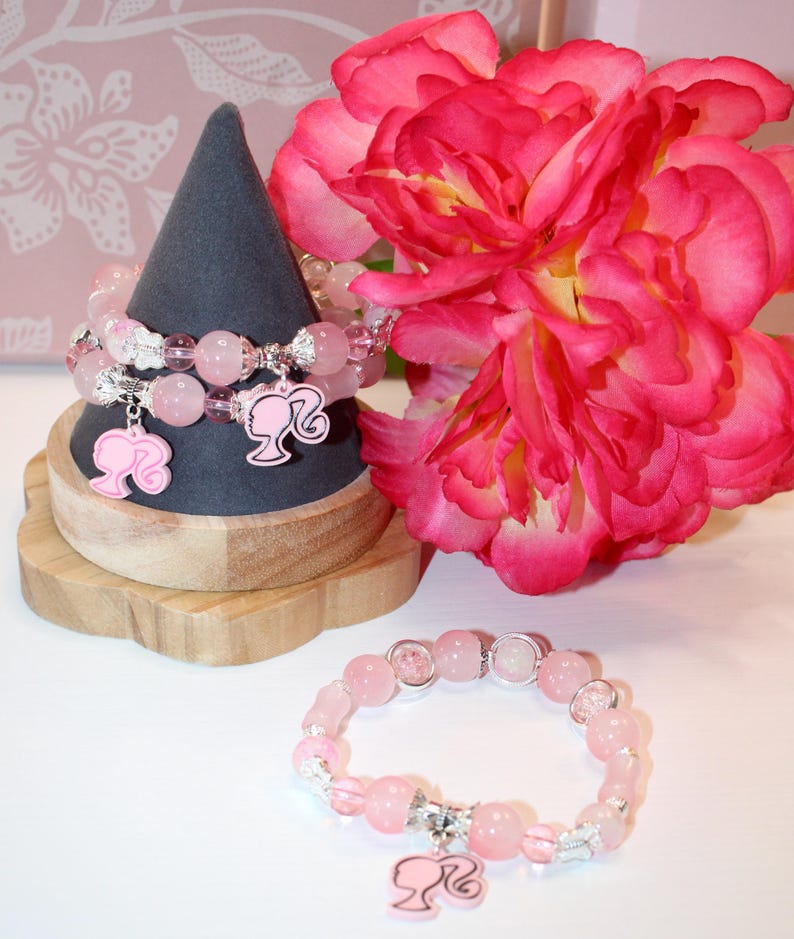 Op de afbeelding: Roze kralenarmbanden met zilveren accenten en Barbie silhouet bedels worden getoond op een houten standaard. Op de achtergrond een grote, levendige roze bloem. De armbanden hebben lichtroze kralen.