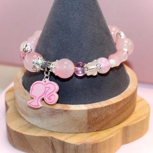Op de afbeelding: Roze kralenarmband met zilveren accenten en een roze bedel met een silhouet. De armband wordt gepresenteerd op een grijze kegelvormige standaard op een houten basis. De kralen zijn van verschillende maten en vormen.