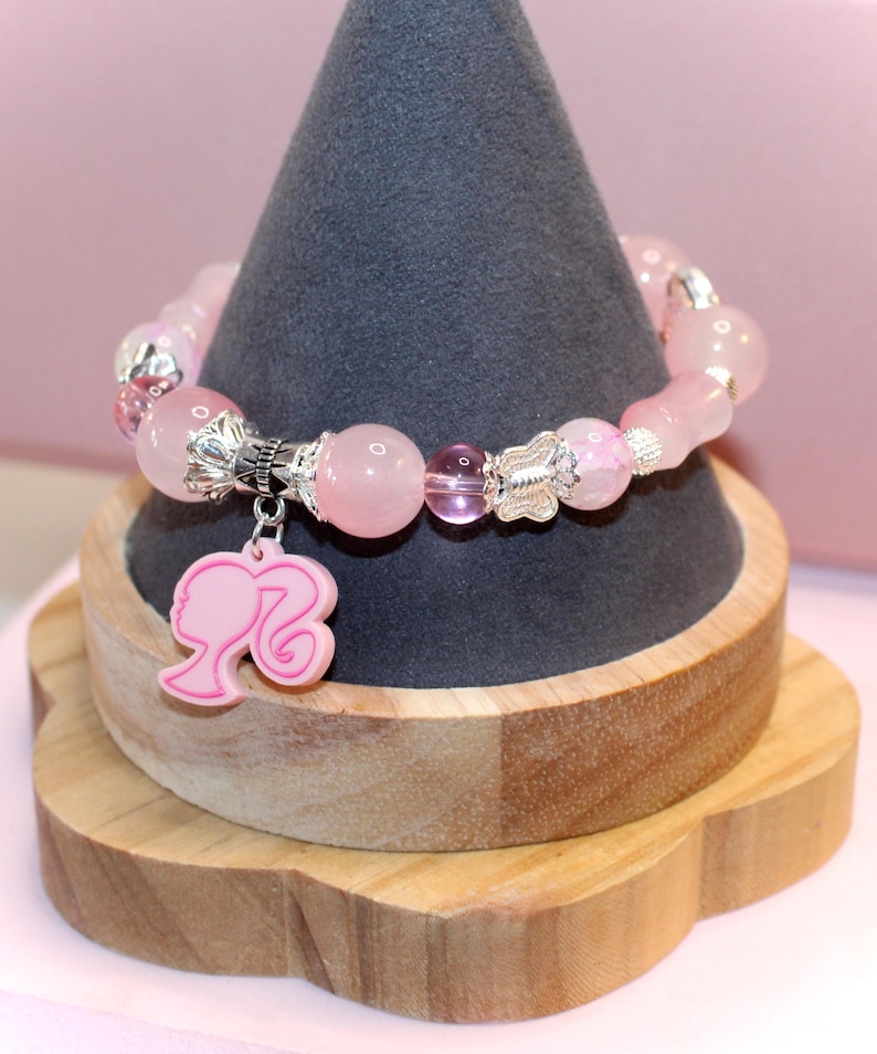 Op de afbeelding: Roze kralenarmband met zilveren accenten en een roze bedel met een silhouet. De armband wordt gepresenteerd op een grijze kegelvormige standaard op een houten basis. De kralen zijn van verschillende maten en vormen.