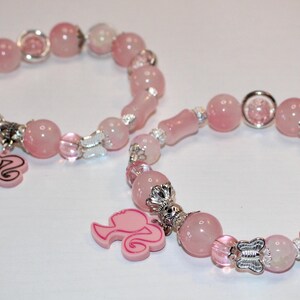 Op de afbeelding: Twee roze kralenarmbanden met zilveren accenten en bedels. De armbanden hebben ronde en langwerpige kralen, met bedels van een silhouet. De armbanden liggen op een wit oppervlak.