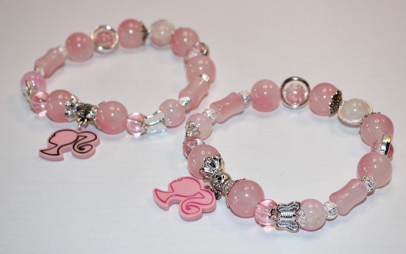 Op de afbeelding: Twee roze kralenarmbanden met zilveren accenten en bedels. De armbanden hebben ronde en langwerpige kralen, met bedels van een silhouet. De armbanden liggen op een wit oppervlak.