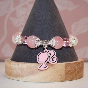 Op de afbeelding: Een roze kralenarmband met een zilveren bedel met een silhouet van een vrouwenprofiel. De armband heeft roze en transparante kralen en wordt gepresenteerd op een grijze kegelvormige standaard.