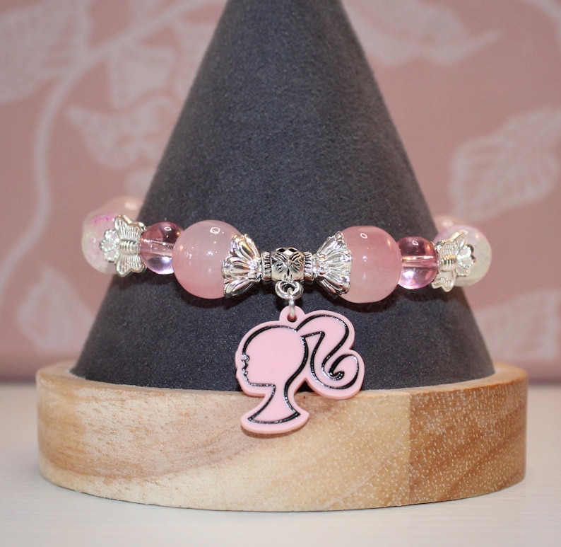 Op de afbeelding: Een roze kralenarmband met een zilveren bedel met een silhouet van een vrouwenprofiel. De armband heeft roze en transparante kralen en wordt gepresenteerd op een grijze kegelvormige standaard.