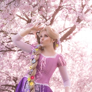Könnte beinhalten: Eine Person in einem lila-rosa Kleid mit einem langen, geflochtenen blonden Haar, das mit bunten Blumen geschmückt ist. Im Hintergrund ein blühender Kirschbaum. Das Kleid hat ein Korsett-Oberteil und einen fließenden Rock.