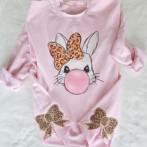 May include: Pink sweatshirt with a white bunny wearing a leopard print bow and blowing a bubble. The sweatshirt has leopard print bows on the cuffs.