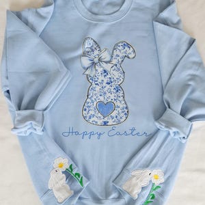 May include: A light blue sweatshirt with a blue and white floral bunny design and the text "Happy Easter". The sleeves have white bunny appliques with a white flower.