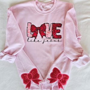 May include: Pink sweatshirt with a red sequined bow and the words "LOVE like JESUS" on the front. The sleeves have red sequined bows on the cuffs.