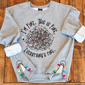 May include: A gray sweatshirt with the text "I'M FINE. THIS IS FINE. EVERYTHING'S FINE." above a tangled string of colorful Christmas lights. The cuffs feature embroidered lights. The sleeves are lined with white fleece.