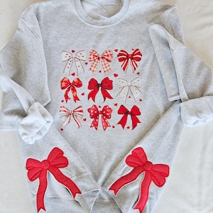 May include: A gray sweatshirt with red bows printed on the front and red bow patches on the sleeves.