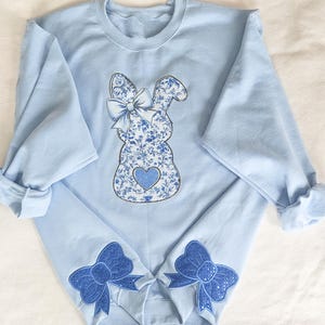May include: A light blue sweatshirt with a blue and white floral bunny applique on the front. The bunny has a blue bow and a heart on its chest. The sweatshirt has blue bows on the cuffs.