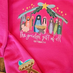 May include: Hot pink sweatshirt with a graphic of the Nativity scene, including the Holy Family and the Three Wise Men. The text "the greatest gift of all" and Matthew 1:21 are printed below the image. A smaller graphic of a manger is on the sleeve.