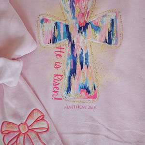 May include: Pink sweatshirt featuring a colorful cross design with gold glitter accents. The cross is painted with blue, pink, and white brushstrokes. Text reads "He is Risen!" and "Matthew 28:6". A pink bow is visible on the sleeve.