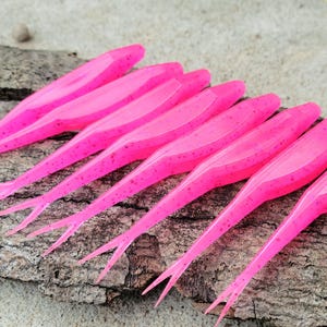 May include: Bright pink soft plastic fishing lures with a slender, elongated body and a forked tail. The lures are arranged in a row on a piece of weathered wood. The lures are approximately 4 inches long.