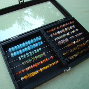 May include: A black display case with a clear lid, showcasing strands of colorful glass beads. The beads vary in size, shape, and color, including blue, green, orange, and yellow. The case is open, revealing multiple rows of beads.