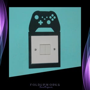 May include: A black light switch cover with a cutout of a video game controller. The switch cover is mounted on a light blue wall.
