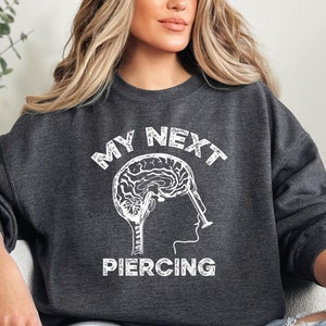 May include: A dark gray sweatshirt with a white graphic of a brain with a piercing needle going through it. The text "My Next Piercing" is above the graphic.