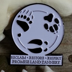 May include: A white enamel pin with black paw prints of a bear, dog, and deer. The pin has a black border and the text "RECLAIM • RESTORE • RESPIRIT PROMISE LAND TANNERY" on a black rectangle below the circle.