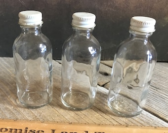 Könnte beinhalten: Drei klare Glasflaschen mit weißen Metallkappen. Die Flaschen sind zylindrisch mit leicht strukturierter Oberfläche. Ein Holzlineal mit dem Text "Promise Land Tannery" befindet sich im Vordergrund.