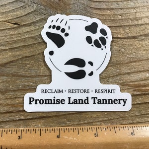 May include: White sticker with black animal footprints in a circle. The text "RECLAIM. RESTORE. RESPIRIT Promise Land Tannery" is below the circle.