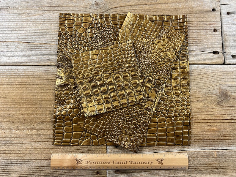 May include: Gold crocodile embossed leather pieces. The leather is textured and has a shiny finish. The pieces are arranged on a wooden surface with a ruler that reads "Promise Land Tannery".
