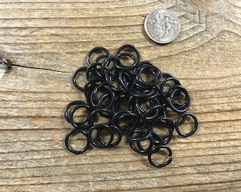 May include: A pile of black jump rings, approximately 1 inch in diameter, on a wooden surface. A US quarter is included for size reference.