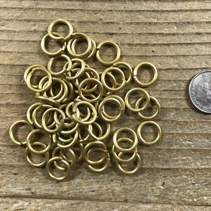 May include: A pile of 50 gold jump rings, approximately 1/2 inch in diameter, on a wooden surface. A US quarter is included for size reference.